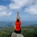 A woman in a red shirt relaxes on a mountain peak with arms raised, enjoying the scenic view.