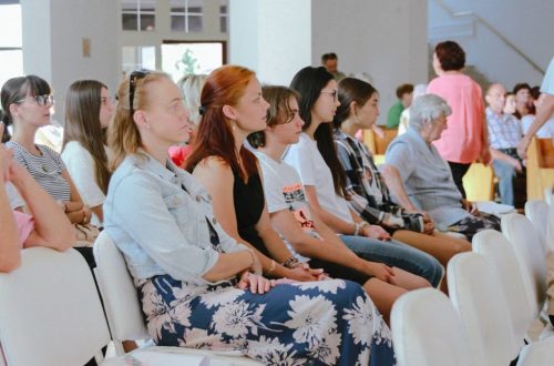 A diverse group of people sitting attentively during an indoor meeting or event, captured in a spacious room.